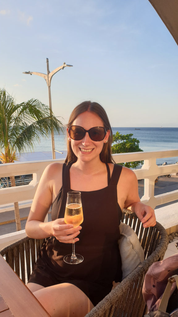 A girl sits at a bar by the coast smiling and holding a glass of bubbly.