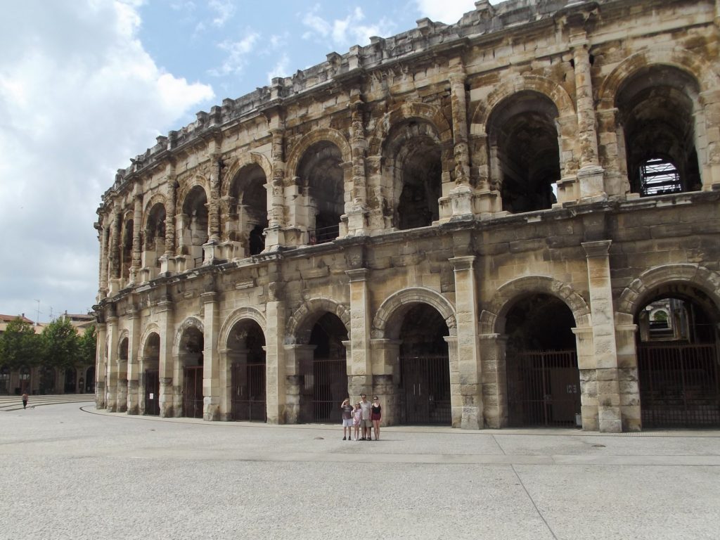 A family stand in front of the impressive Nimes Arena. The archways are tall and intricately carved.