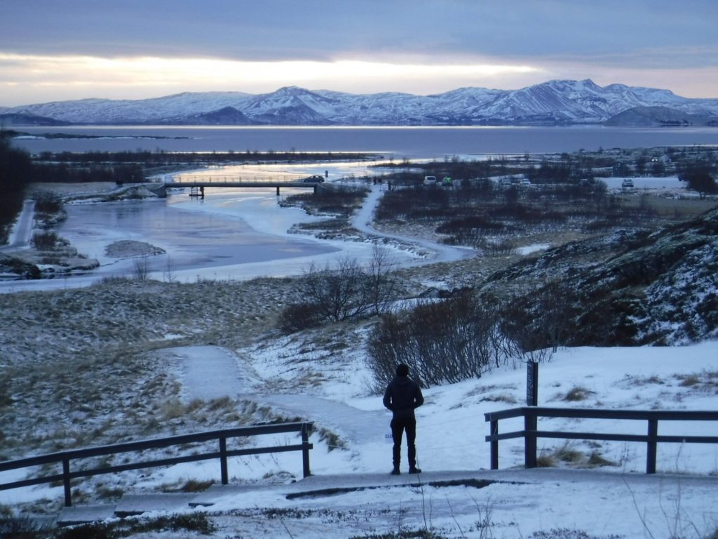 A boy stands looking out at a frozen scene - Thingvellir National Park. A car crosses a small bridge over a shimmering lake.