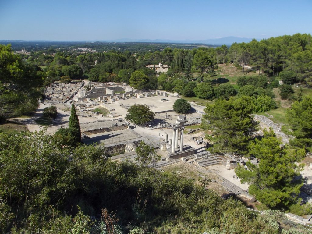 The ancient remains of a roman city, Site Archéologique de Glanum shows off straight roads, columns and walls. Trees and green fields surround the site.