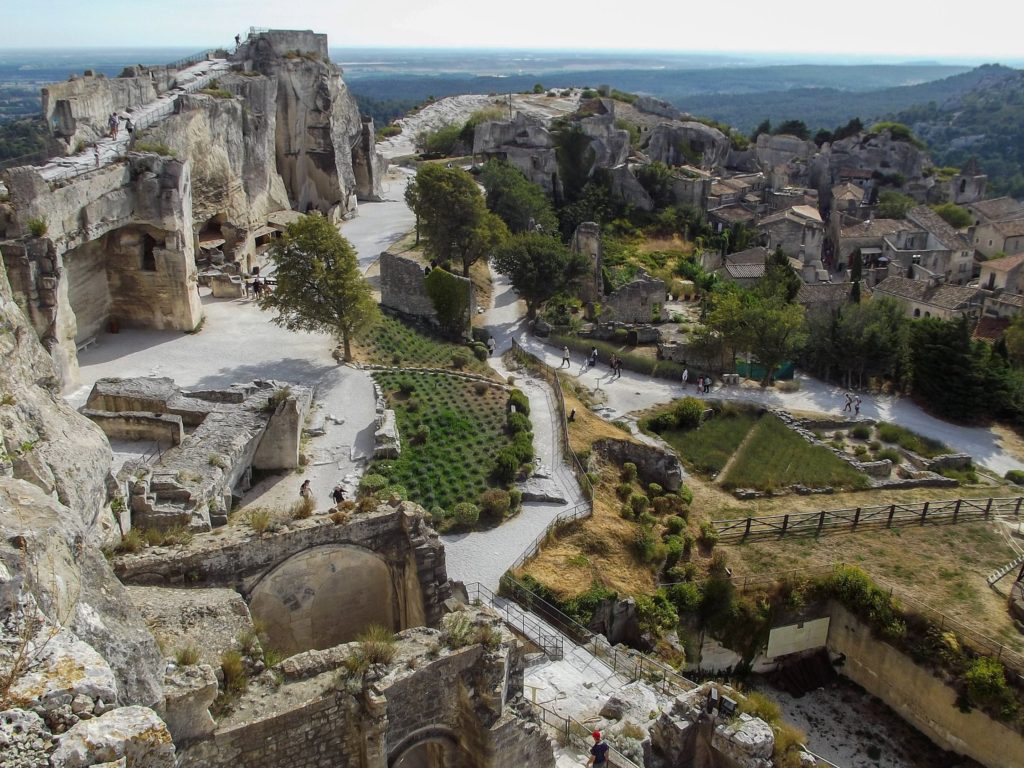 The impressively tall remains of Château des Baux extend for quite some distance. The surrounding area is inhabited and is surrounded by greenery.