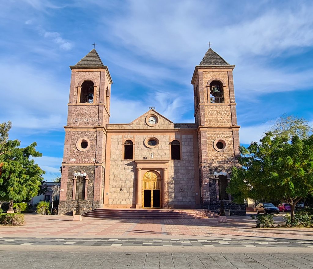 A pink and orange stone cathedral stands in the centre of La Paz. It is neighboured by greenery on both sides.