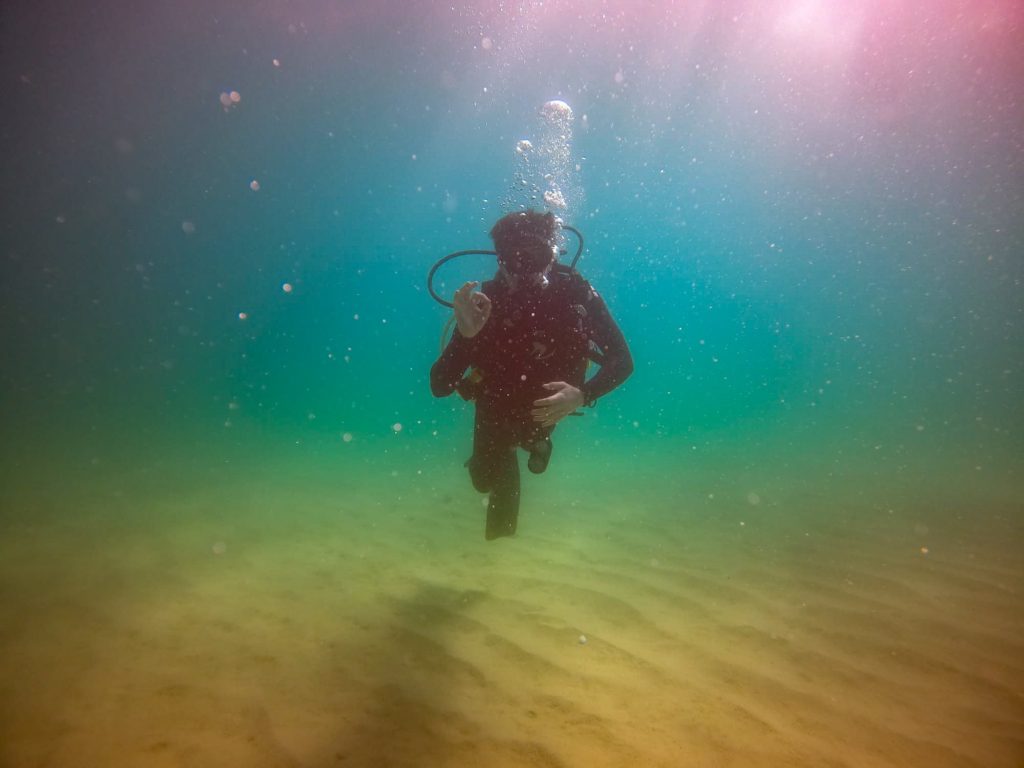 A view underneath the waves: a SCUBA diver gives the "OK" sign. The water is murky and colourful, spanning the whole spectrum of the rainbow.