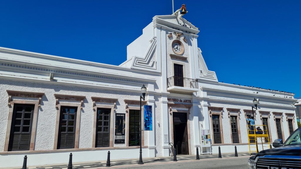 The clean white facade of the Baja California Sur Art Museum stretches almost the whole length of the street. The building is topped with a central clock and a bell.