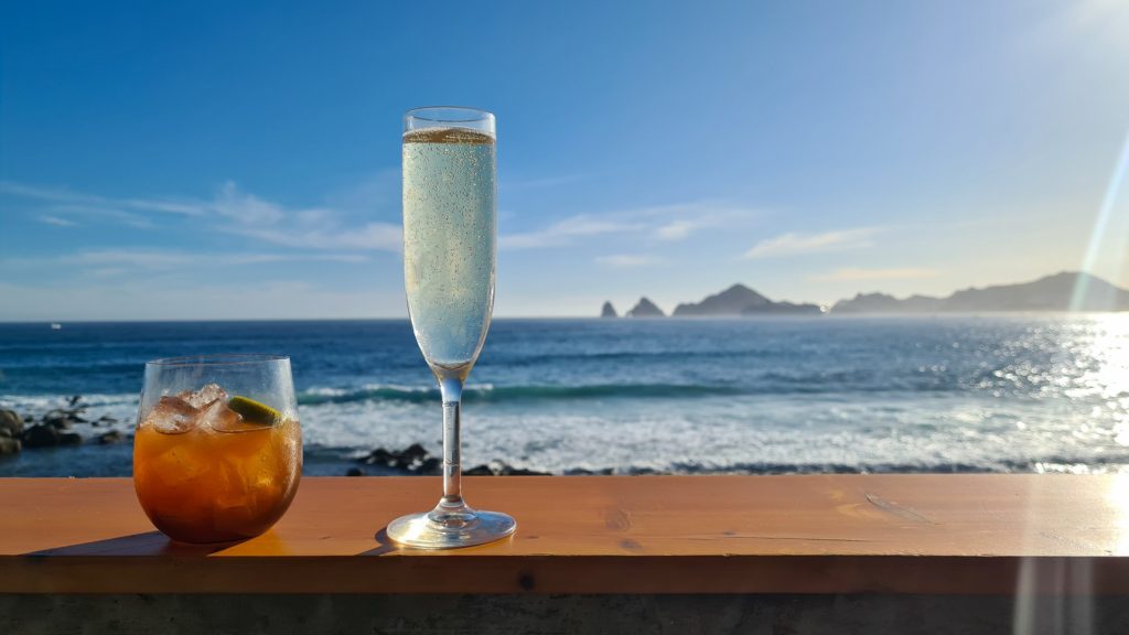 Two cold cocktails on a balcony overlooking the bay in Cabo. The sun reflects off the waves and the famous Arch of Cabo can be seen in the distance.
