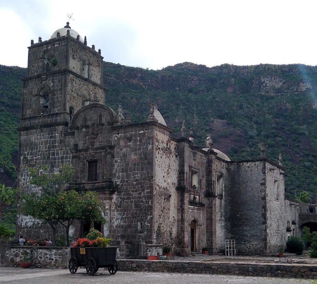 An impressive stone church stands in front of a back-drop of greenery. A tall fruit tree stands at the church entrance.