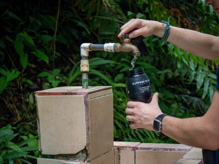 Dan fills up his reusable water bottle at an outdoor fountain. The area is surrounded by greenery.