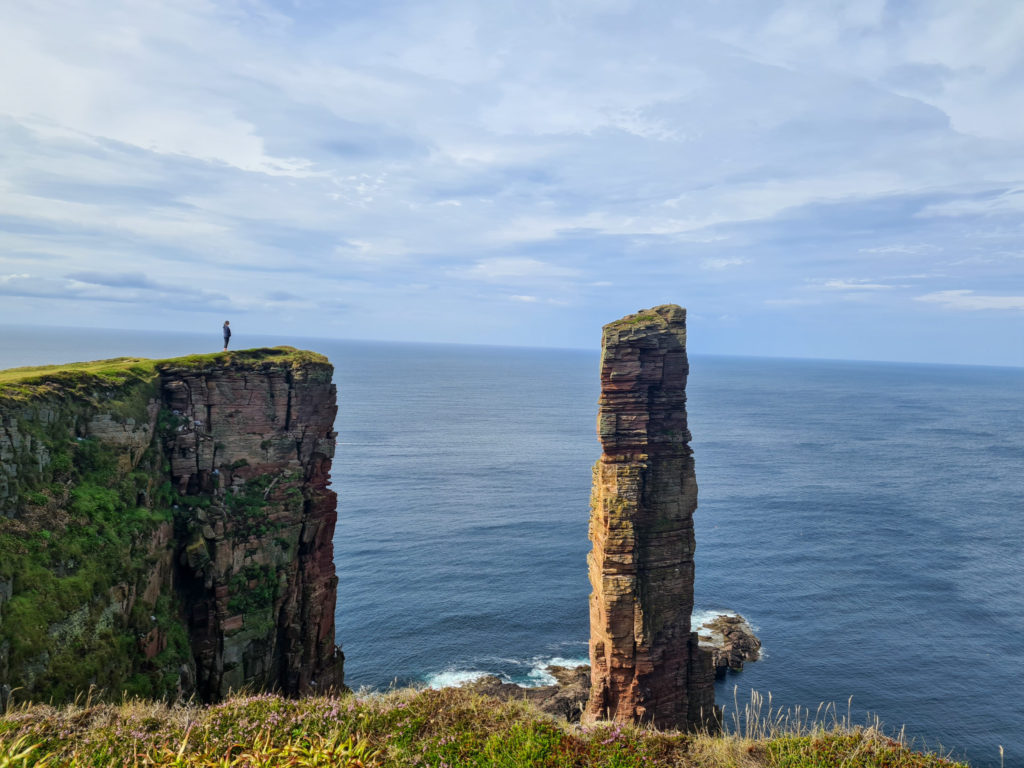 A girl stands alone by the edge of a cliff and gazes at a large sea stack.