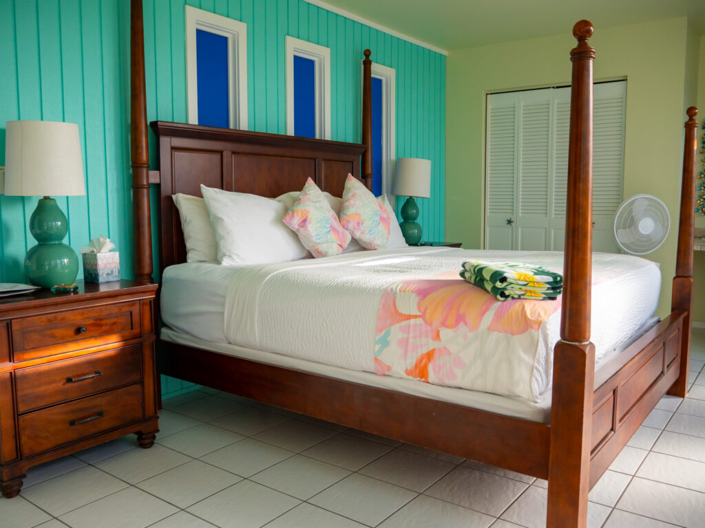 An impressive wooden four-poster bed stands on clean white tiles. A standing fan can be seen in the corner.
