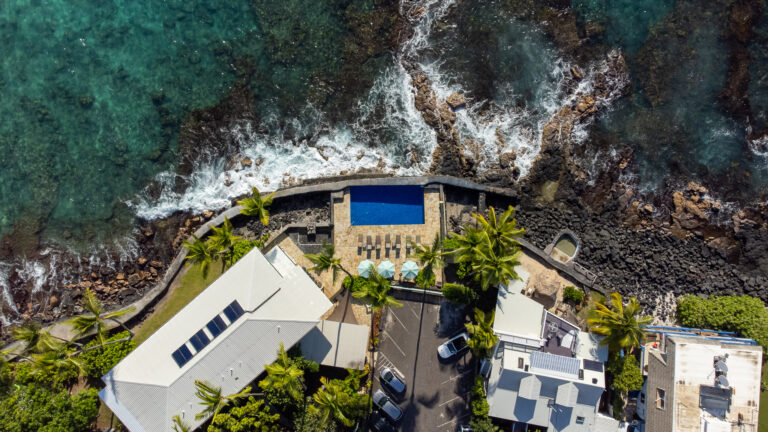 A drone shot of Kona Tiki Hotel. Waves are crashing against the se wall right next to the pool. Greenery is dotted throughout the scene. The water is clear and blue.