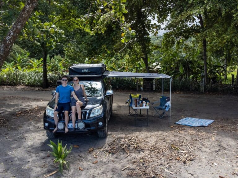 Dan and Lucy sit on the hood of their hired Nomad America camper rental 4x4 car. They are parked on a sandy beach under the shade of palm trees. The awning is set up and there is a camping table and chairs ready for breakfast outside.