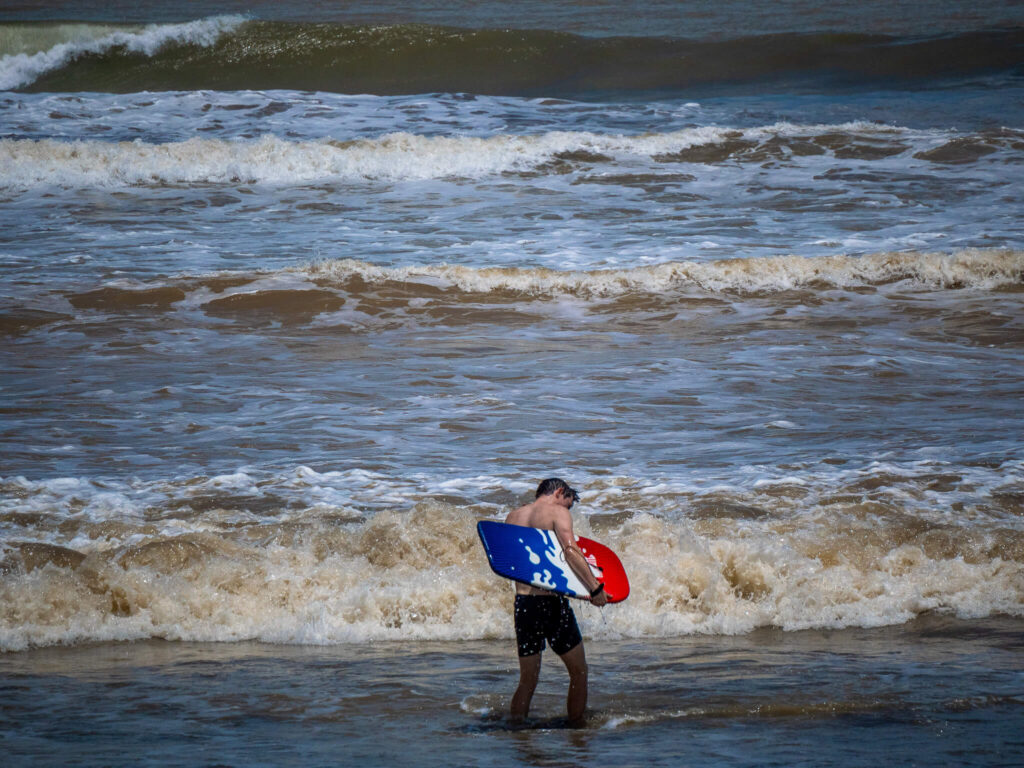 Dan steps into the sea carrying a bodyboard under his arm.