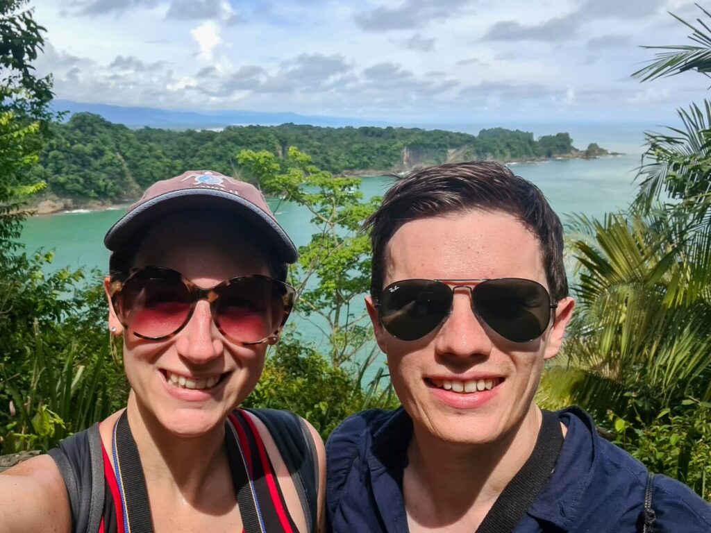 Dan and Lucy smiling as they stand in an opening in the trees with a view of the sea.