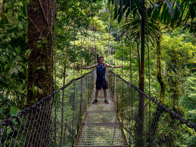 Dan stands on a hanging bridge in the forest, his arms outstretched to his sides.