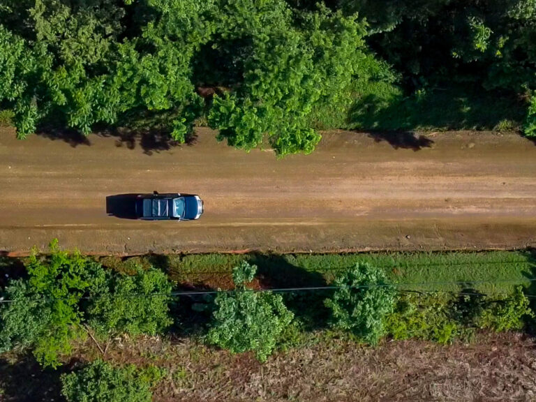 A top-down drone shot of a car driving down a dirt road lined by green trees.