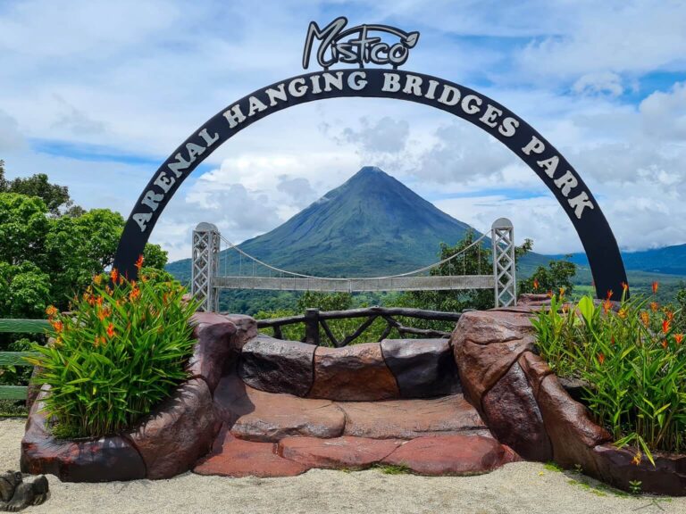 A sign welcoming visitors to Mistico Arenal Hanging Bridges Park. The sign is framed perfectly above a stunning view of Arenal Volcano.