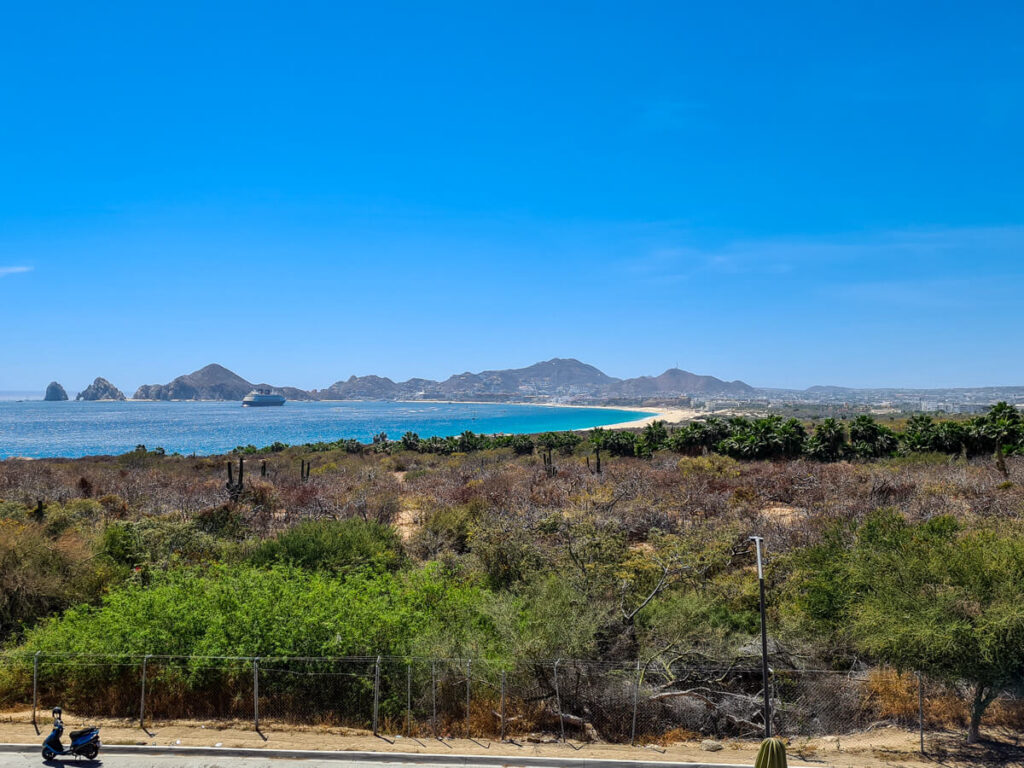 Cabo Bay from a distance. Behind green-brown shrubbery, a curving bay housing a large cruise ship in bright blue waters.