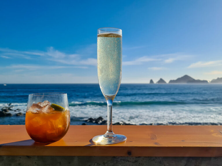 Two cocktails on a seaside bar in the sunshine. Past the drinks is a view of El Arco (The Cabo Arch) across the water.
