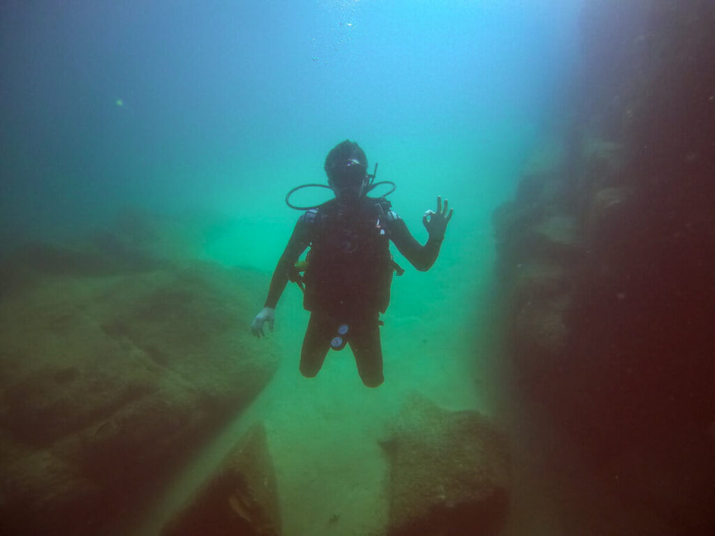 Dan making the OK symbol with his hand, SCUBA diving in Cabo, surrounded by misty green waters.