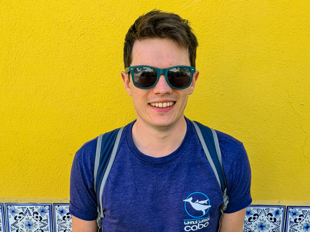 Dan smiling in sunglasses, stood against a bright yellow wall.