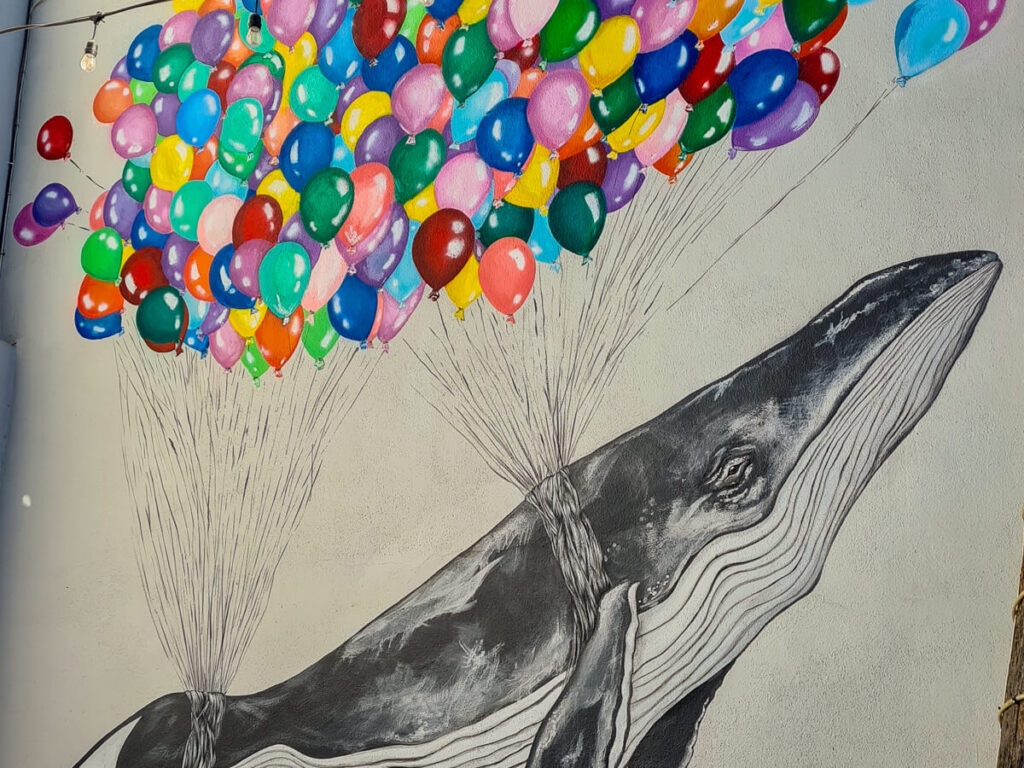 Street art of a humpback whale being held up by lots of colourful balloons.