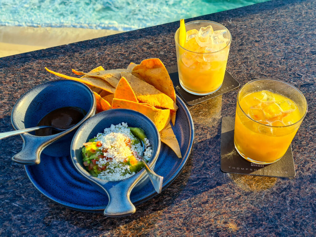 A plate of freshly made guacamole and corn totopos beside two orange cocktails.