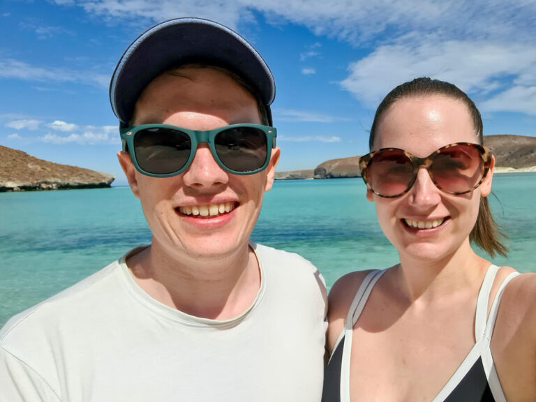 Dan and Lucy smiling in sunglasses, and behind them, a view of Balandra Bay in Mexico.