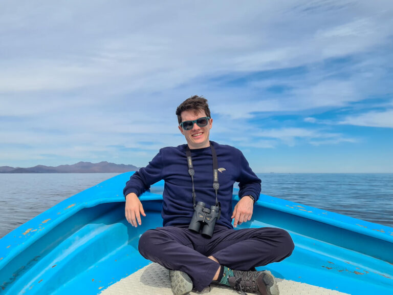 Dan sitting at the front of a small blue boat with his sunglasses on. Mountains can be seen in the distance.