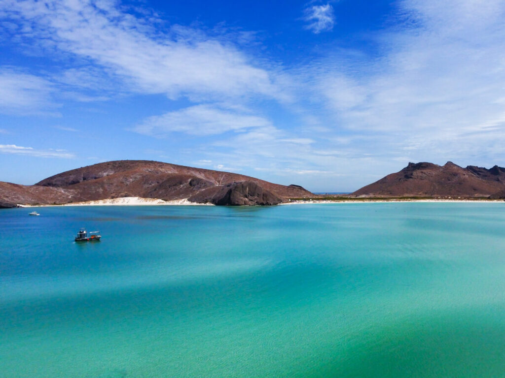 Balandra Bay as viewed from a drone. Red-brown hills surround the bay and two small boats are moving slowly through the calm water.