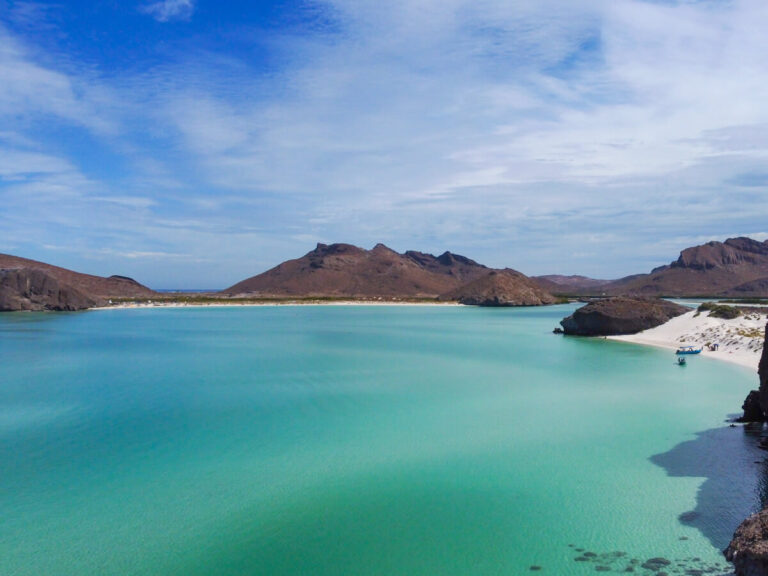 Balandra Bay as viewed from a drone. White sand beaches line the calm waters, and a couple of small boats are sitting anchored in the shallows.