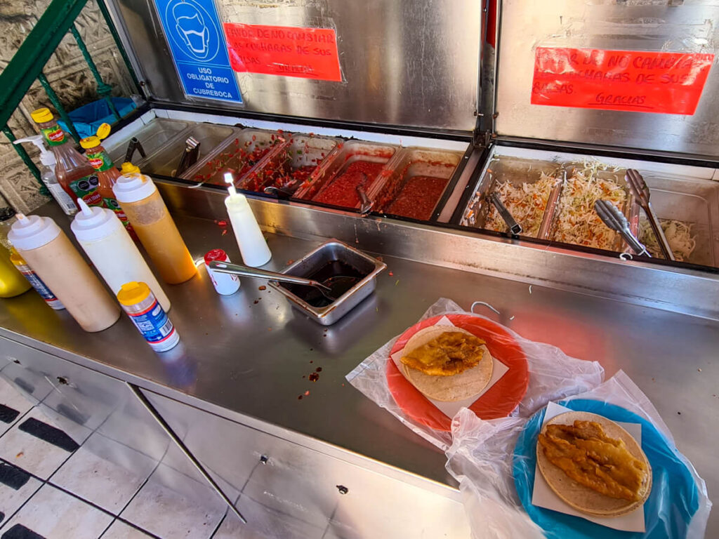 A toppings station at a taco restaurant. There are a variety of sauces to choose from as well as fresh coleslaw and onions.