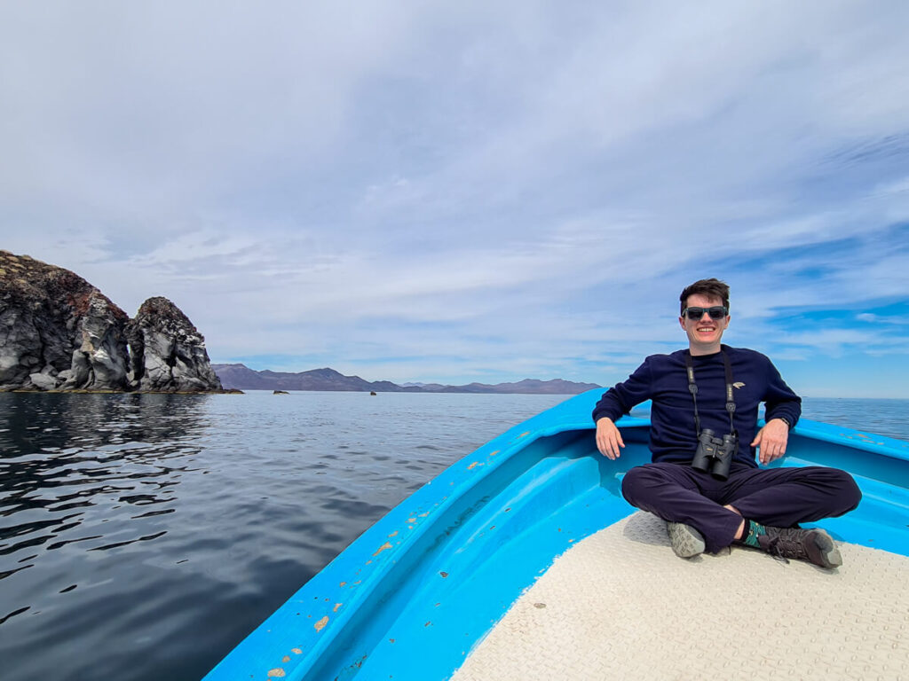 Dan sitting at the end of a small boat in Loreto Bay National Park. He is smiling and is wearing sunglasses and carrying binoculars around his neck.