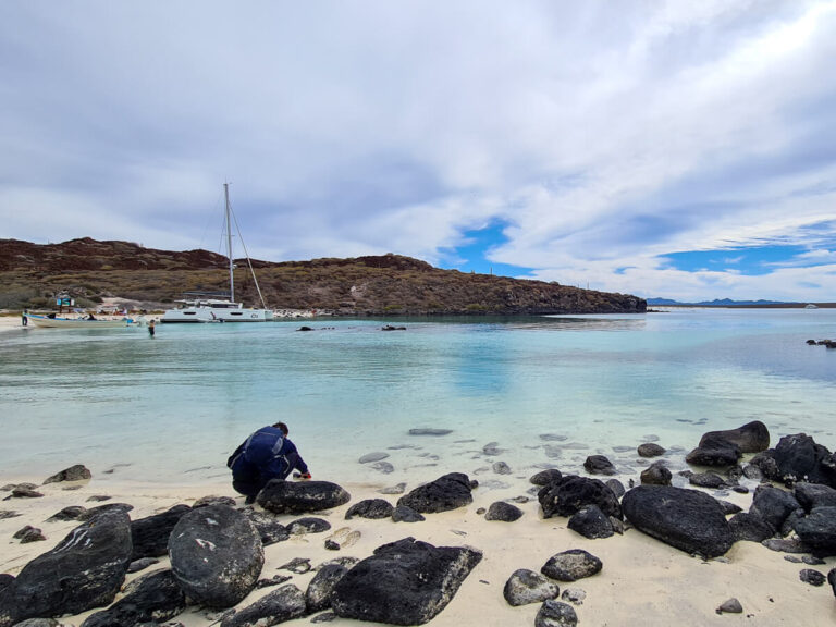 Dan crouching on a beach on Isla Coronado. A couple of boats are anchored nearby and a couple of people are paddling in the water.