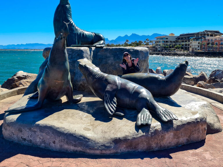 Lucy crouching amongst the sea lion sculptures in Loreto marina. She is putting her hands out as if they were flippers and smiling in the sunshine.