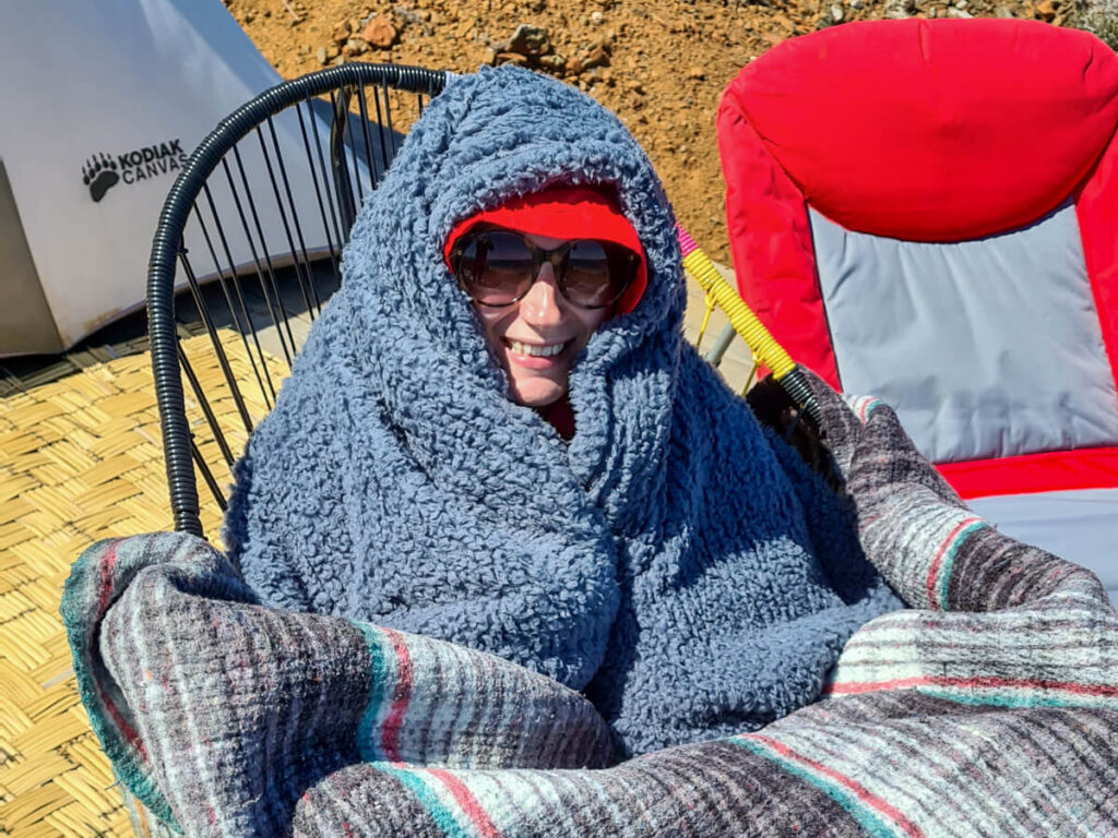 Lucy wrapped up in a bundle of multiple blankets on a deck chair at Magdalena Bay Whales Camp. She is smiling in the sunshine and has on a pair of sunglasses.