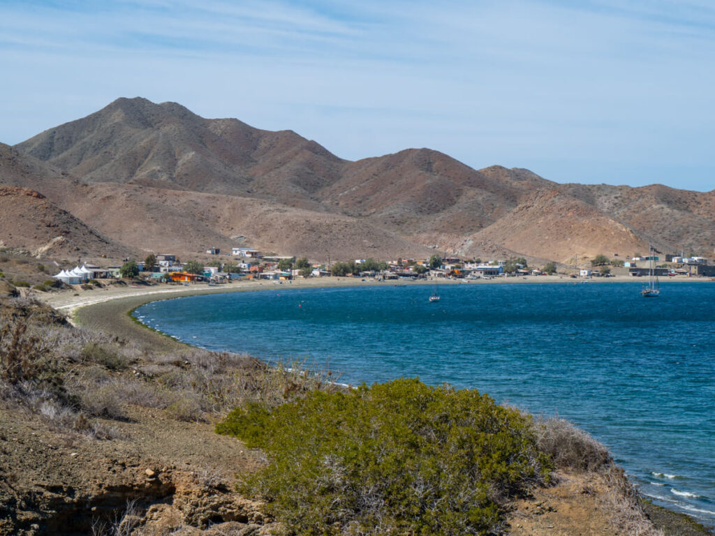 The curving land of Magdalena Bay in the sunshine. In the calm waters there are a couple of small fishing boats.