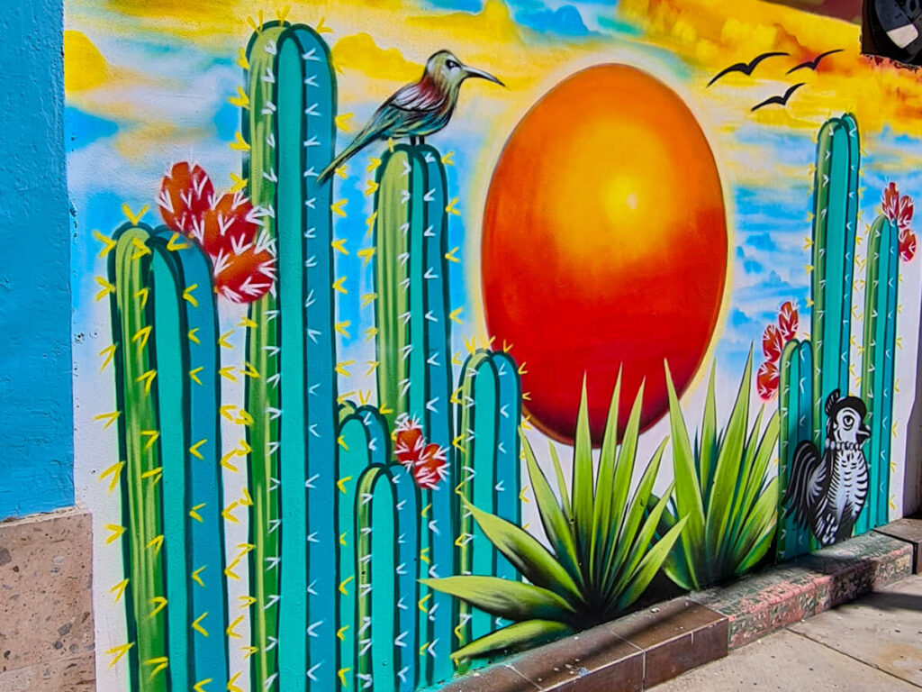 Street art of a bright orange sun and birds among large green cacti on a wall in La Paz, Mexico.