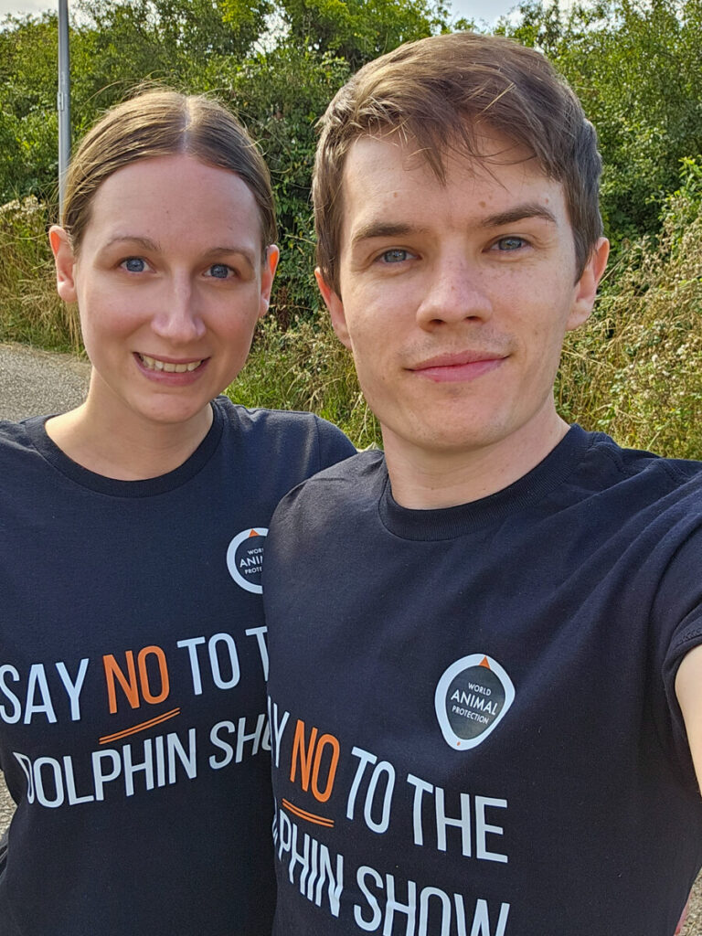 Dan and Lucy standing next to each other with green bushes behind them. They are wearing matching shirts which read "World Animal Protection. Say no to the dolphin show". These photo was taken in support of a campaign to stop the practise of keeping dolphins in captivity so as to get them to perform for an audience.