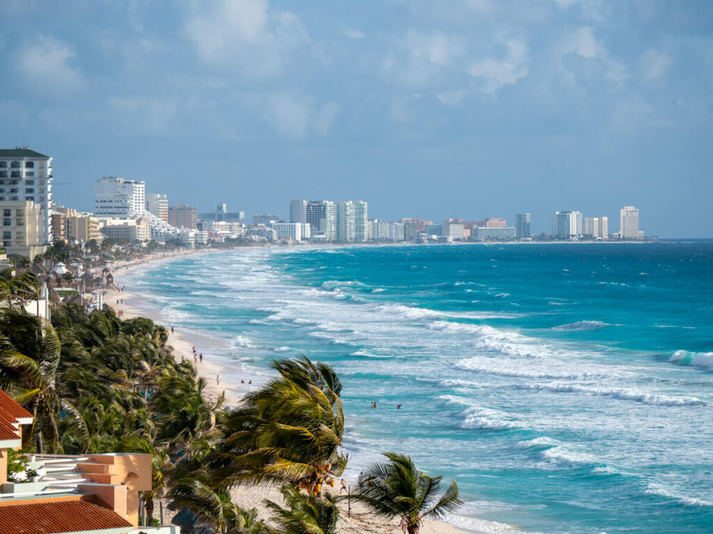 The sprawling Cancún Hotel Zone covered in high-rise resorts with turquoise waves crashing on the shore and palm trees swaying in the wind, leading into the distance.