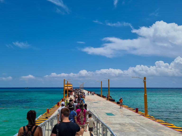 A queue of people waiting for the Cozumel Ferry on a long concrete pier in Playa del Carmen, surrounded by bright blue waters.