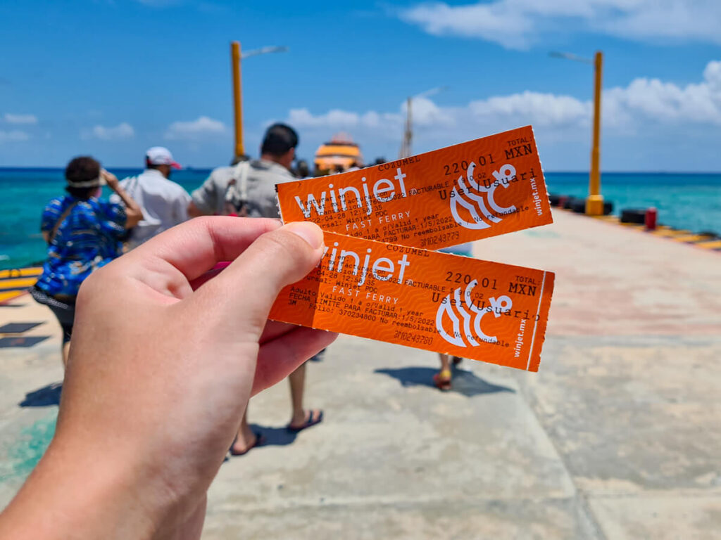 A hand holding two orange tickets for the Winjet Cozumel Ferry in front of a concrete dock.