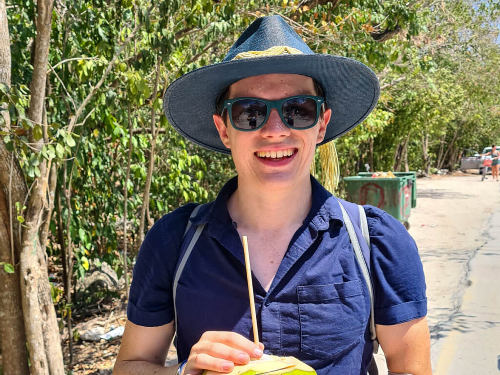Dan in a wide brimmed hat and sunglasses holding a coconut in Tulum, Mexico.