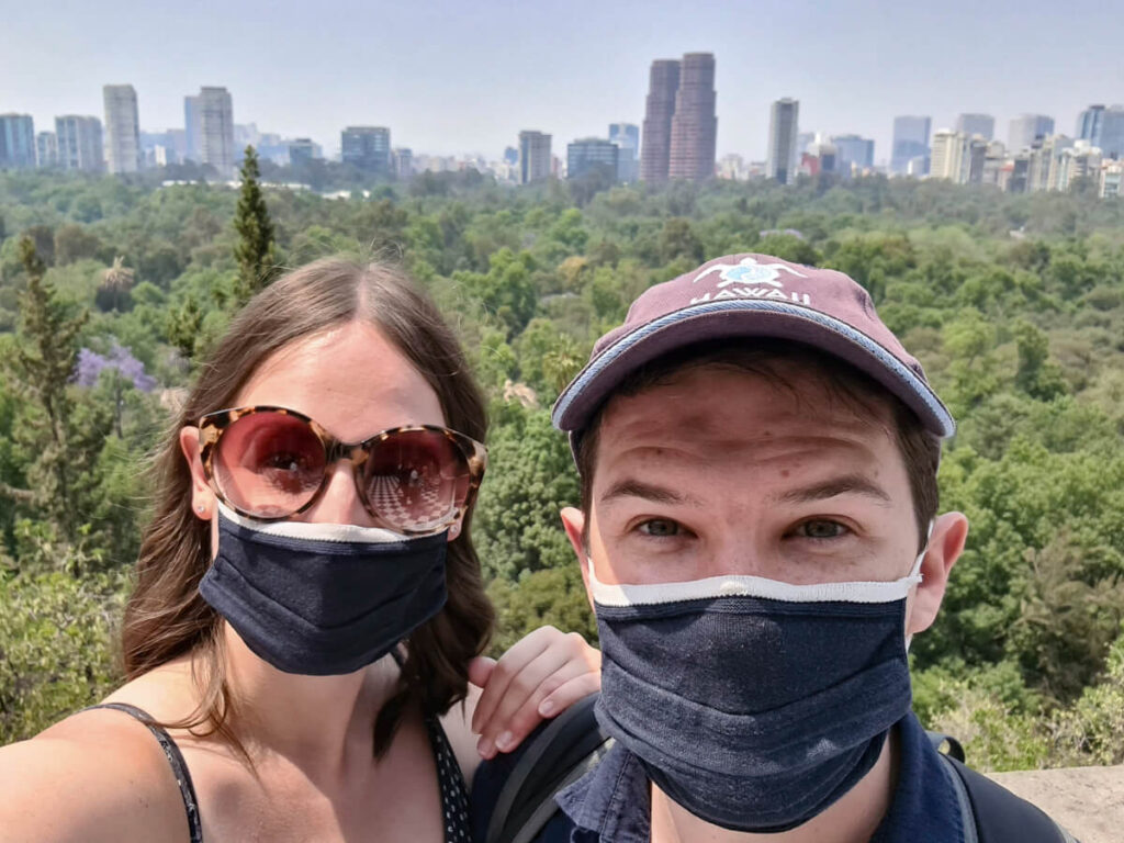 Lucy and Dan wearing face-masks. A park and then the tall buildings of Mexico City are behind them.