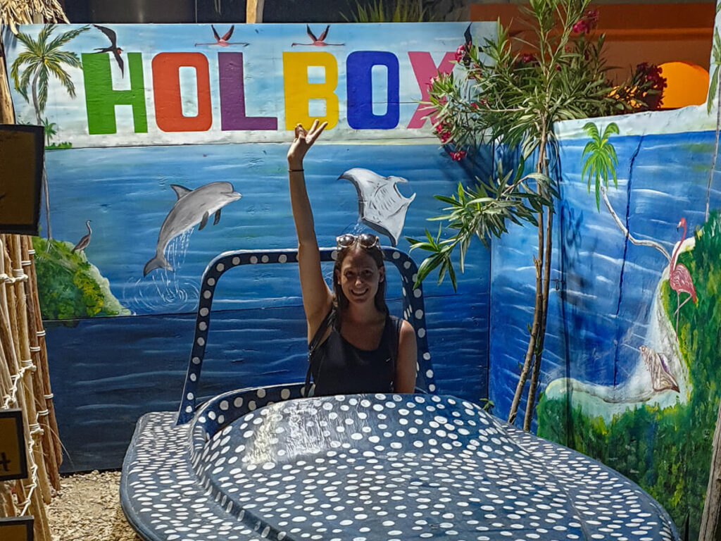 Lucy sitting in a small boat painted like a whale shark in front of a colourful sign that spells Holbox.