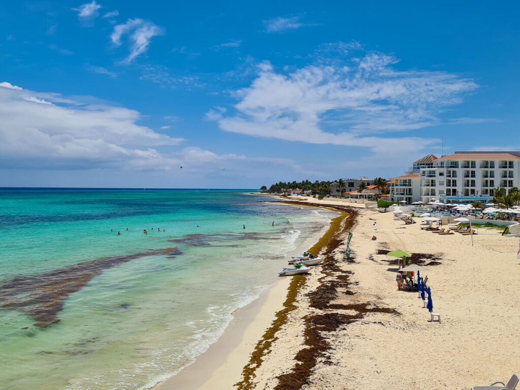 A long sandy beach lined with buildings and leading to the clear blue sea on the coast of Playa del Carmen.
