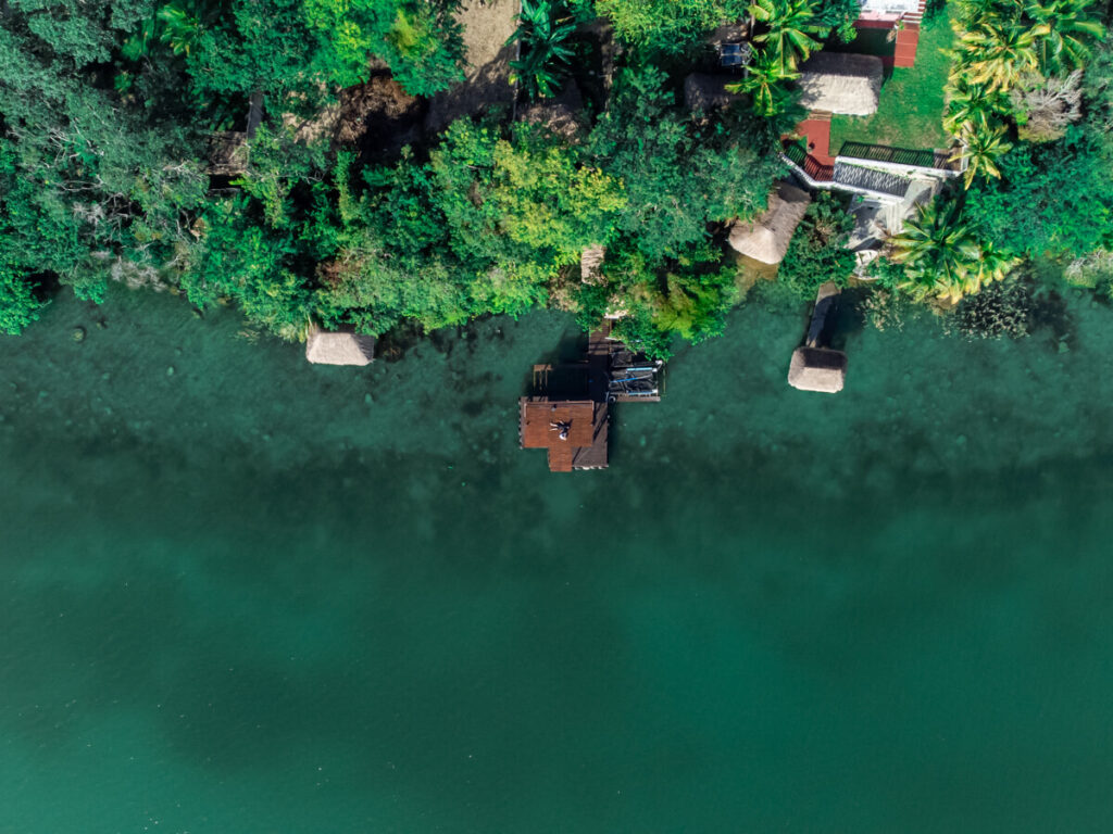 A section of Bacalar Lagoon as viewed from a drone high above the trees. The Eco-Hotel Casa Lamat is mostly hidden beneath the surrounding greenery. Lucy and Dan are relaxing on a wooden dock.