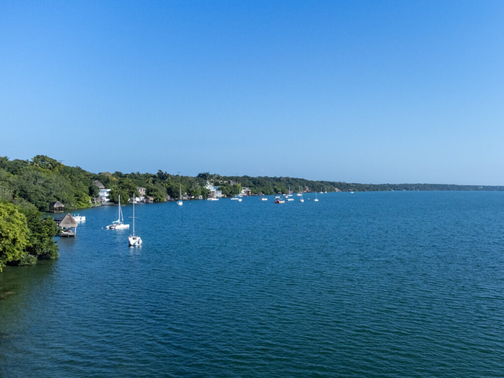 Sailboats on Bacalar Lagoon in the sunshine float along the shore, which is lined with private docks belonging to hotels in Bacalar.