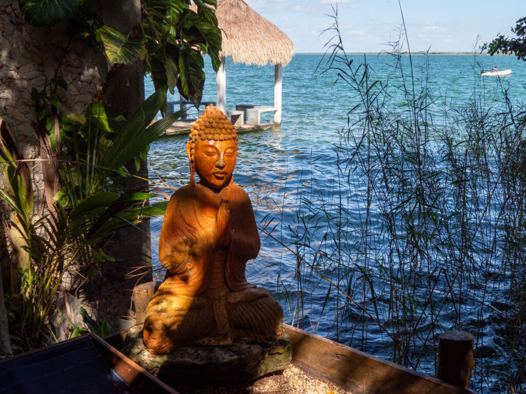 A full body sculpture of the Buddha in a seated position. The sculpture has been placed on the edge of a dock on the vast and beautiful Bacalar lagoon.