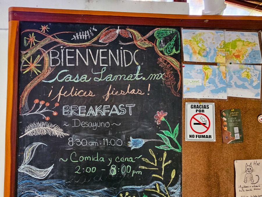 The Casa Lamat noticeboard has the opening times for the restaurant, a map for guests to pin where they have come from, and a "no smoking" sign.