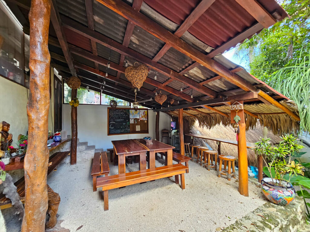 The Casa Lamat restaurant - a small selection of outside seating including a large table with benches and a long tall table with accompanying stools. Wooden carvings and decorations hang from the makeshift roof.